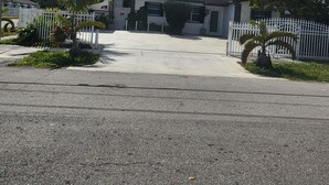 A residential property with a paved driveway leading to a house with a white exterior and a small porch. There are palm trees and a white fence surrounding the property. - Cozy 1-bedroom studio in pleasant hollywood with AC (West Park)