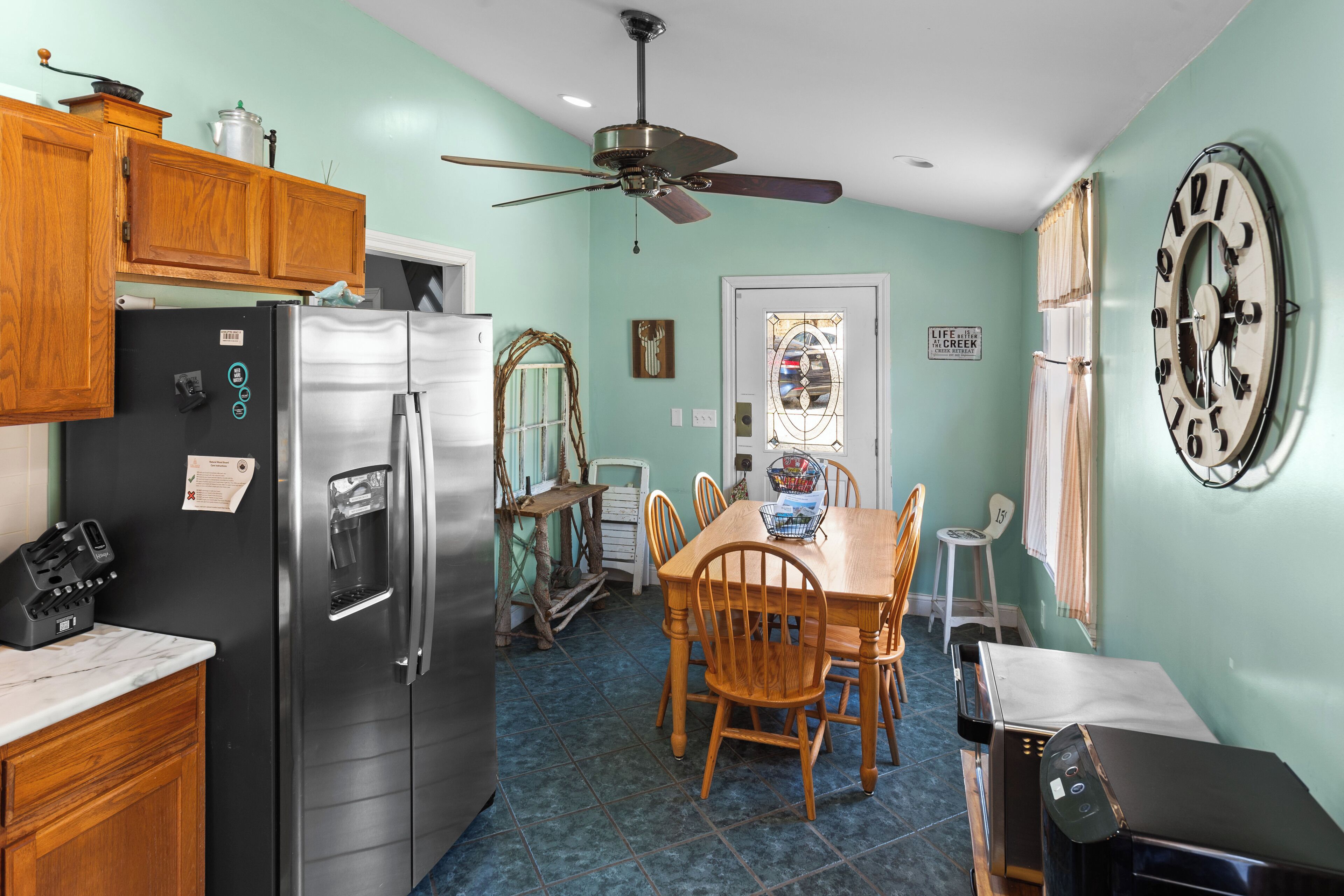 A kitchen with wooden cabinets, a stainless steel refrigerator, a dining table with chairs, and a wall clock.