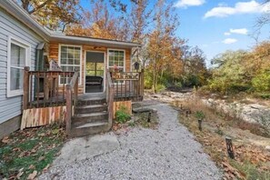 A wooden cabin with a porch, a gravel path leading to the entrance, and autumn foliage in the background. - Private setting between flowing creeks, minutes from Cincinnati attractions. (Cincinnati)
