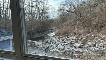 A window view of a rocky stream with bare trees, binoculars, and a tripod on a wooden surface.