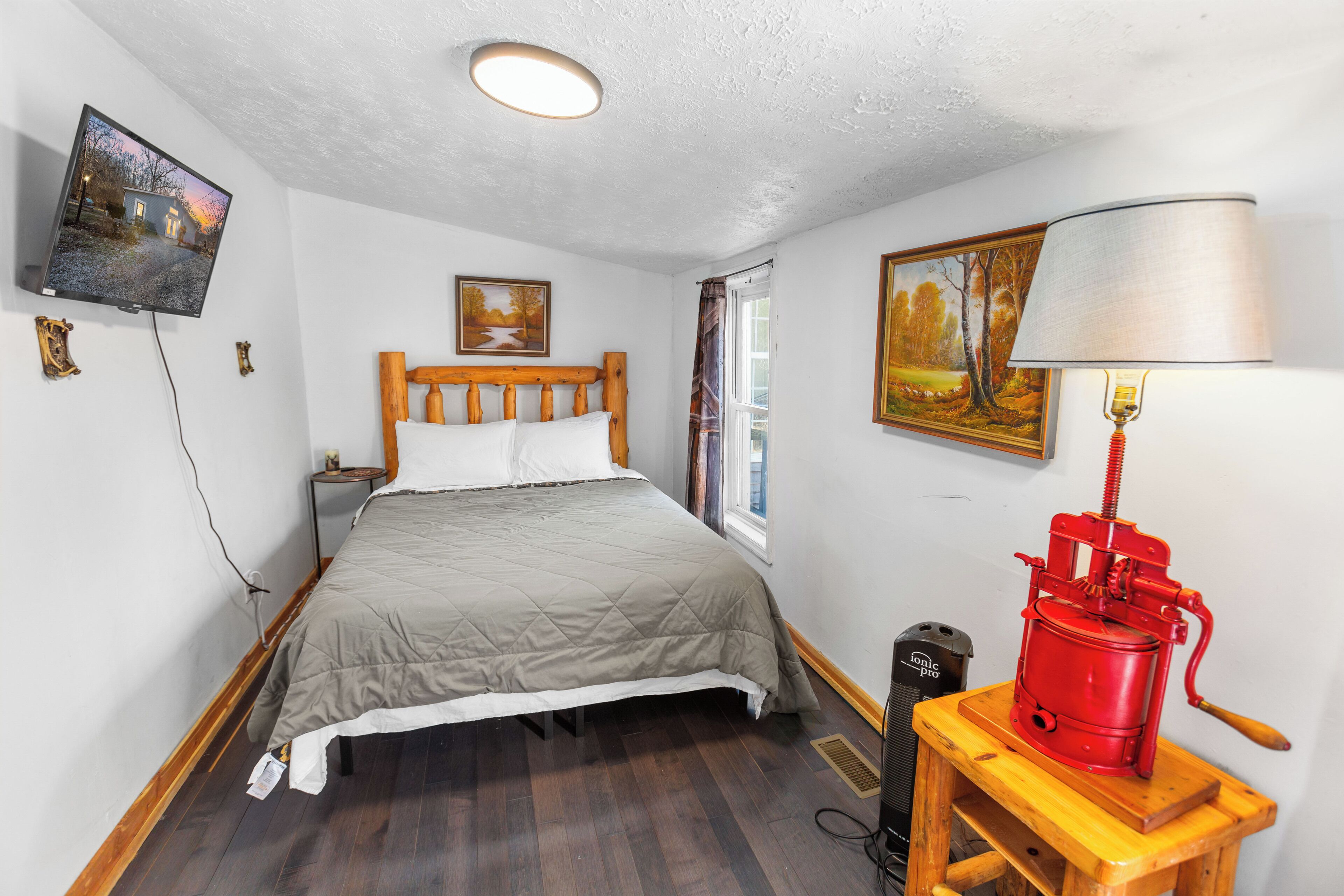 A bedroom with a bed, a wooden headboard, a television mounted on the wall, a window with curtains, and a red appliance on a wooden table.