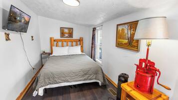 A bedroom with a bed, a wooden headboard, a television mounted on the wall, a window with curtains, and a red appliance on a wooden table.