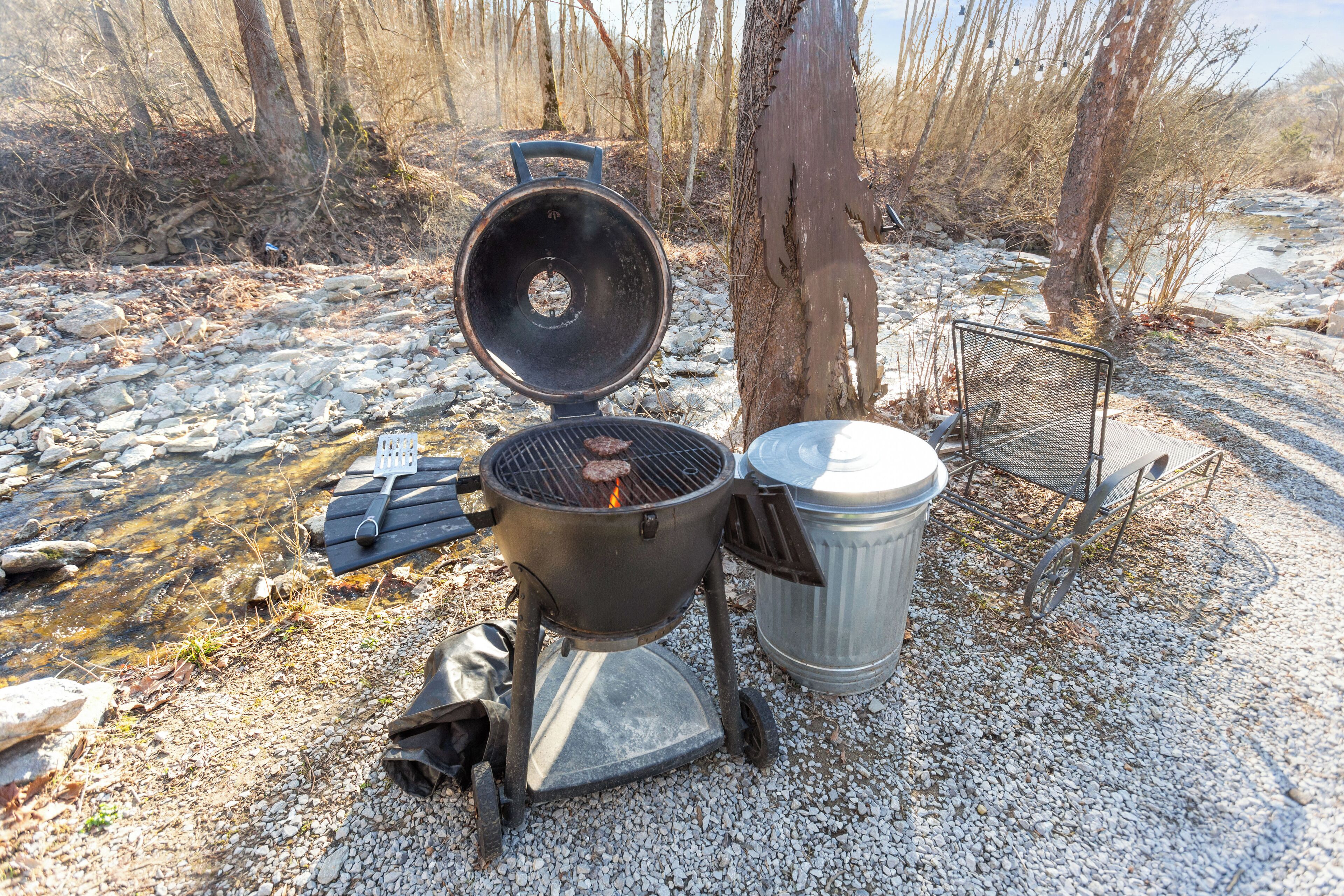 A portable grill with a lid open, a trash can, and a metal chair.