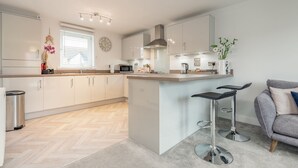 A modern kitchen with white cabinets, a central island, bar stools, a microwave, and a window with blinds. - Belmont House | Kingsbarns (Kingsbarns)
