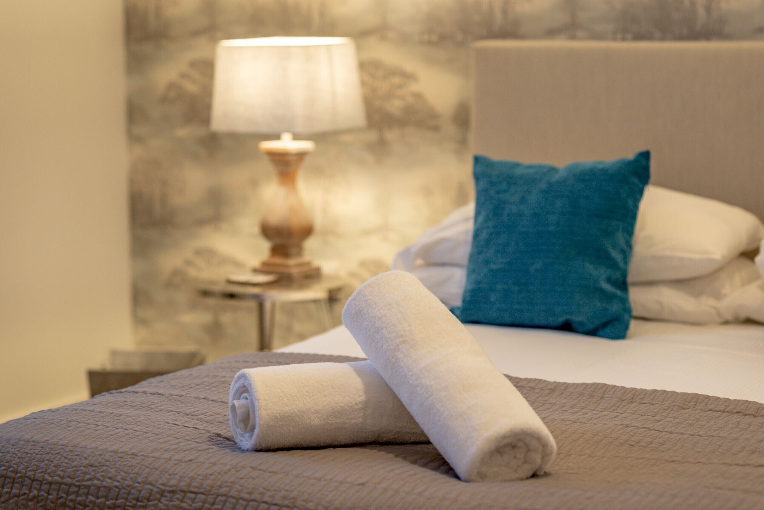 A neatly made bed with a rolled white towel, a blue pillow, and a white pillow on a textured bedspread.