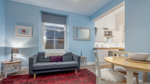 A living room with a grey sofa, a red patterned rug, a wooden dining table with a white bowl, and a kitchen in the background. - Little Haven | St Andrews Town Centre (St Andrews)