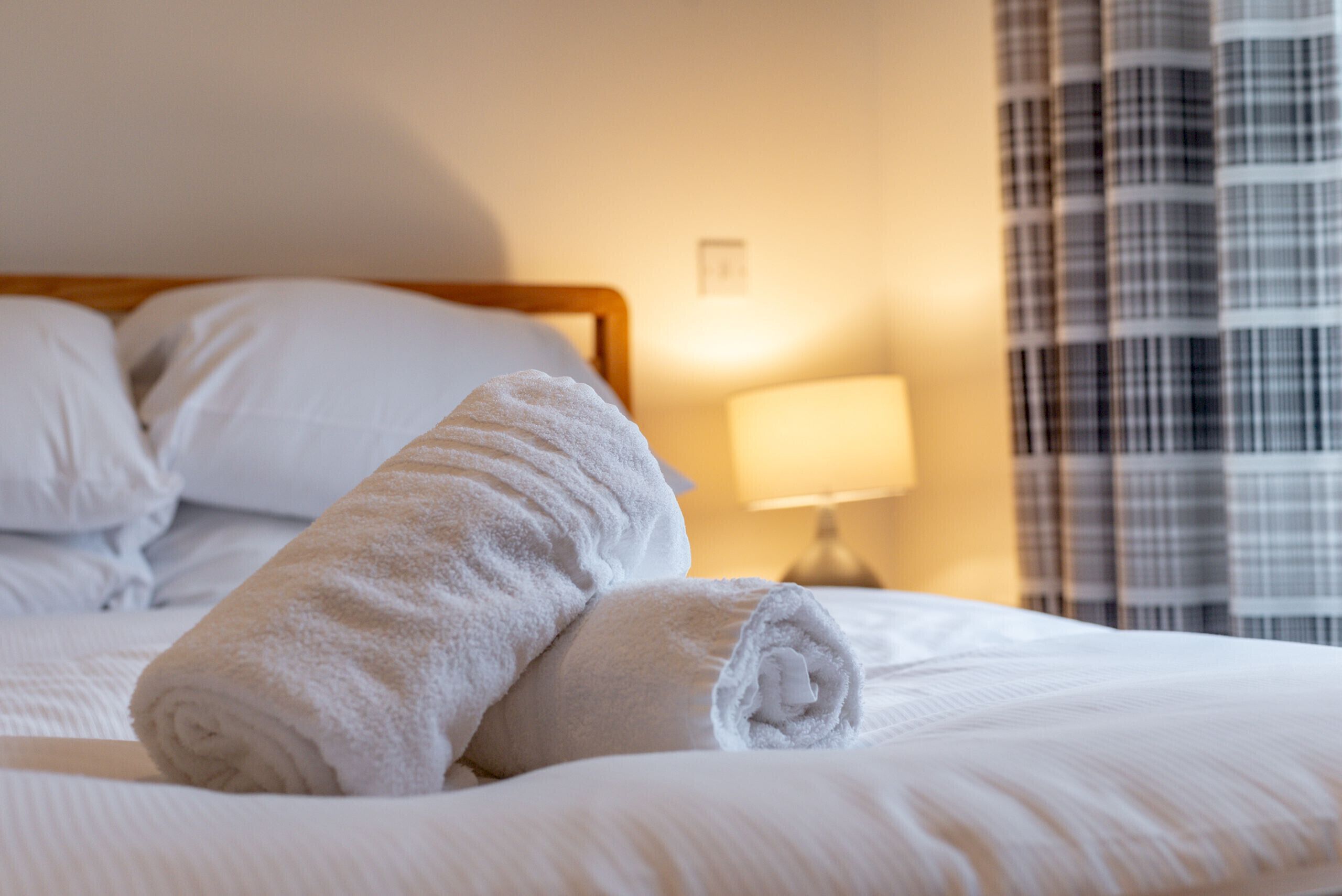 Un lit bien fait, avec du linge de lit blanc et des serviettes pliées, une lampe de chevet et une fenêtre ornée d’un rideau à carreaux.