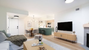 A modern living room with a sofa, coffee table, and a TV. There is a dining area with chairs and a kitchen in the background. - True Luxury - Pool, HT, Sauna (Whistler)