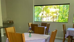 A dining area with wooden chairs and tables covered with checkered tablecloths, a large window with a view of greenery, and a shelf with glassware. - Pousada Conchas Do Mar (Cairu)