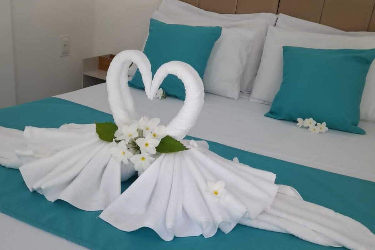 Cama bem arrumada com edredom verde-água, almofadas brancas e um enfeite de cisne feito com toalhas e flores.