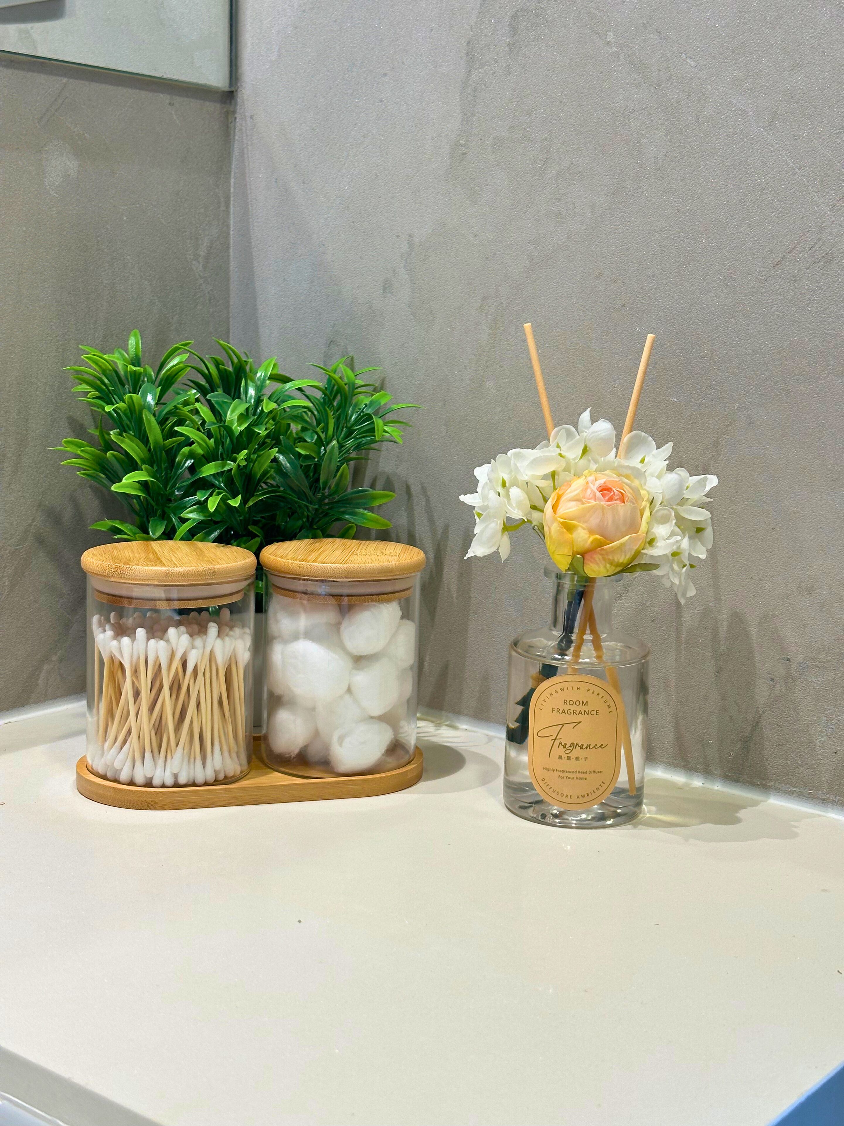 Two glass jars with wooden lids, one containing cotton swabs and the other with white cotton balls, and a small bottle of room fragrance with a label.