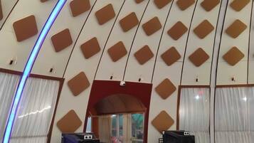 A room with a unique geometric ceiling design, a small seating area with a sofa and chairs, and two large speakers.
