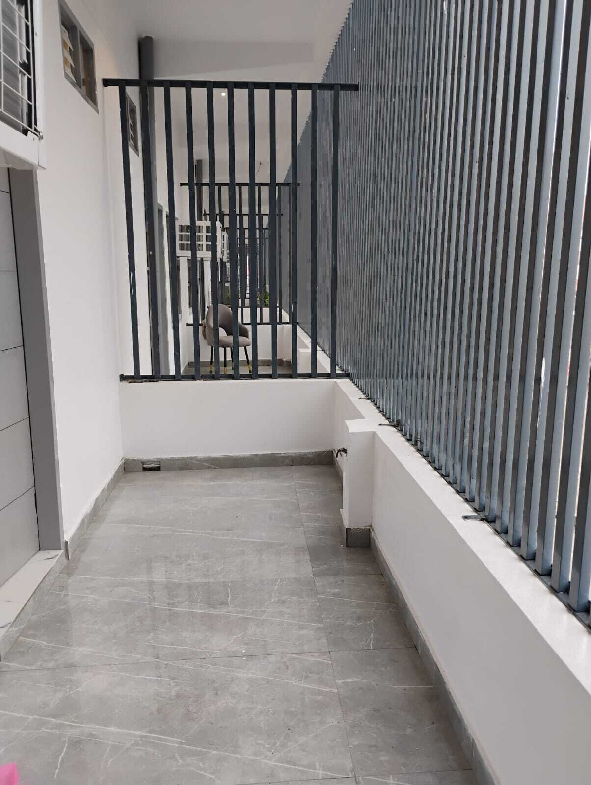 A narrow, tiled balcony with a black metal railing, a chair, and a white trash bin.