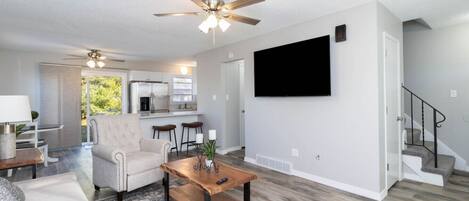 A modern living room with a sofa, coffee table, and ceiling fan. There is a kitchen area with a refrigerator and a TV mounted on the wall.