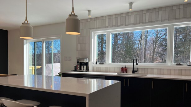 A modern kitchen with a large island, bar stools, a coffee machine, and a view of trees through large windows.