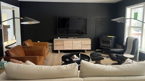 A modern living room with a large flat-screen TV, a ceiling fan, a fireplace, leather sofas, and a dining area with a table and chairs.