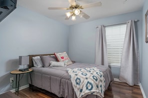 A bedroom with a bed, a bedside table, a lamp, a ceiling fan, and a window with curtains.
