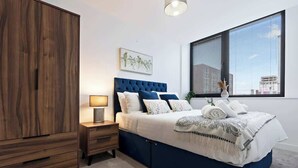 Une chambre avec une grande armoire, un lit avec une literie bleue, une table de chevet avec une lampe et une fenêtre avec des stores.