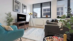 Un salon moderne doté d’un téléviseur à écran plat, d’un canapé, d’une table basse avec des magazines et des verres, et d’une grande fenêtre offrant une vue sur la ville.