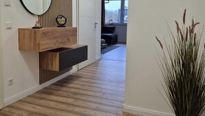Un pasillo con piso de madera, tocador de madera con espejo, planta en maceta y puerta que da a otra habitación.