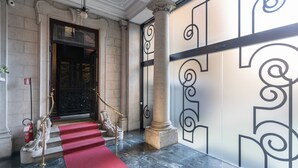 Une entrée imposante, avec un tapis rouge menant à une porte noire, flanquée de colonnes richement décorées et d’un mur orné de motifs géométriques.