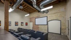 Un salón de conferencias con una gran mesa, varios asientos dispuestos en filas, una pantalla de proyector y una pared de ladrillo.