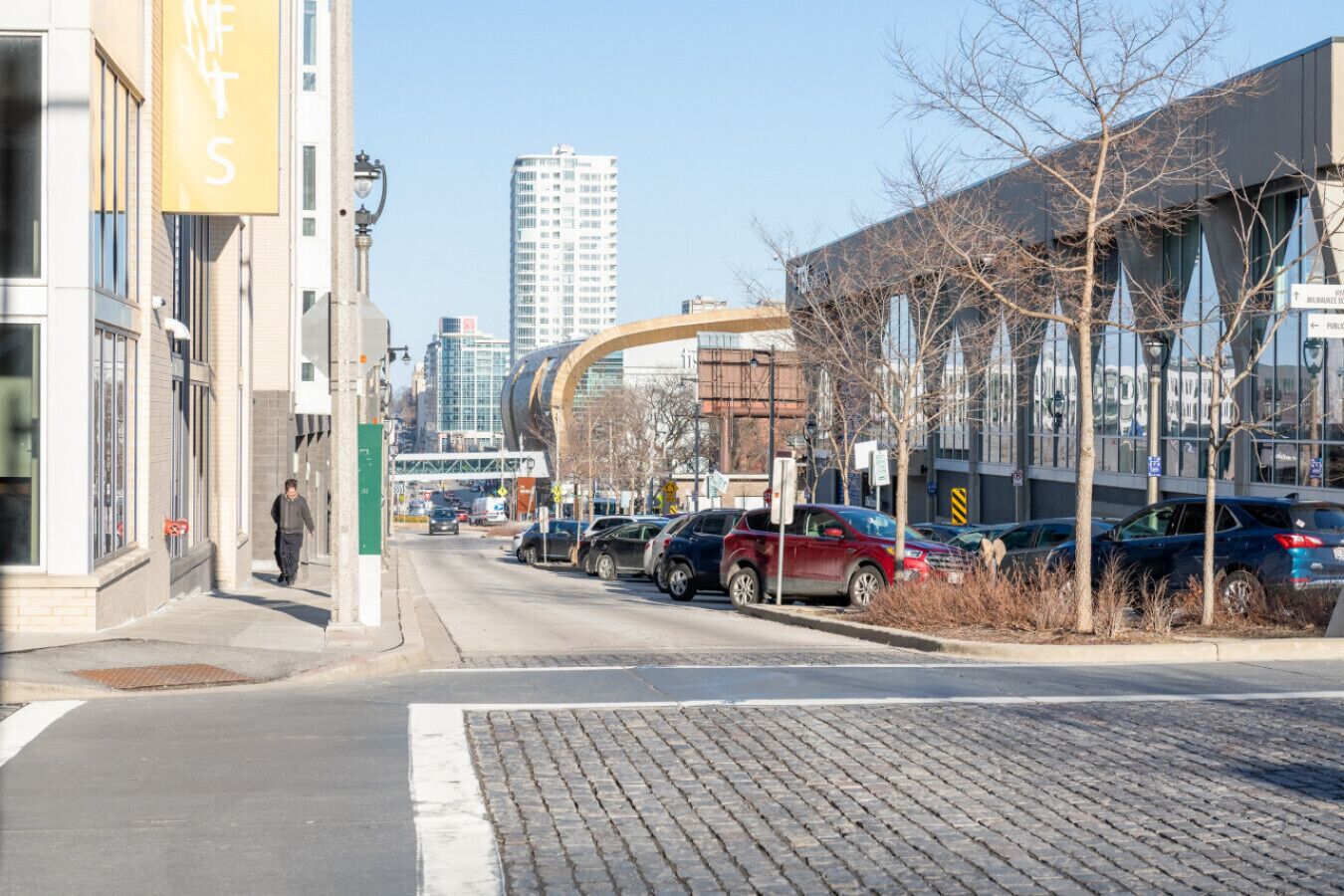 A city street with parked cars, a pedestrian, and modern buildings.