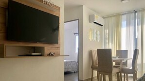 A modern living room with a flat-screen TV mounted on the wall, a dining area with a table and chairs, and a sofa.