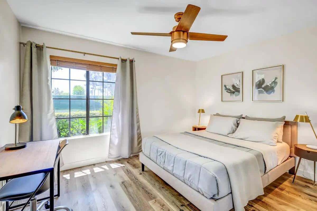A bedroom with a bed, desk, chair, ceiling fan, and two framed pictures on the wall.