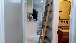 A narrow hallway with a wooden ladder leaning against a wall, leading to a kitchen area with a sink and cabinets.