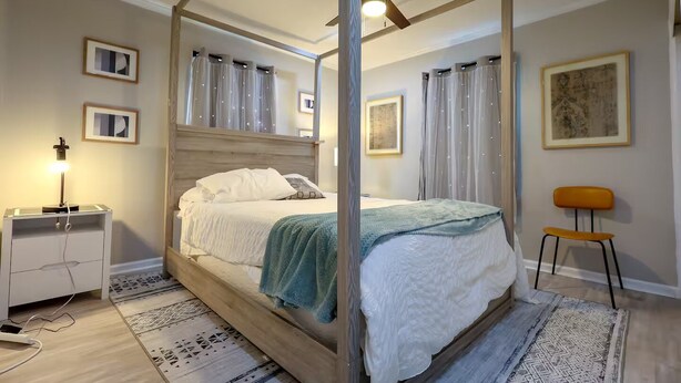 A four-poster canopy bed with a wooden headboard, a bedside table with a lamp, a chair, and a rug.
