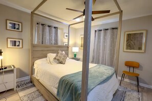 Un lit à baldaquin à quatre poteaux, avec une tête de lit en bois, du linge de lit blanc et un plaid vert. Une table de chevet avec une lampe et une chaise se trouvent sur la droite.