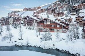 A snow-covered resort with multiple wooden buildings, a river in the foreground, and a mountainous backdrop.