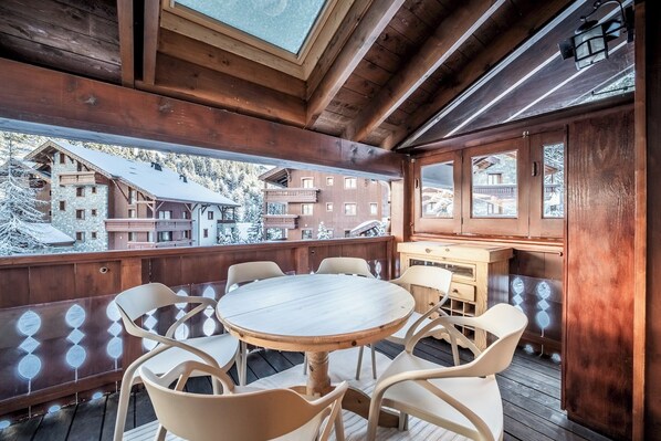 A wooden deck with a round table and chairs, a wooden cabinet, and a view of snow-covered buildings.