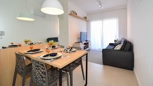A modern dining area with a wooden table set for four, surrounded by chairs. A television is mounted on the wall, and a sofa is visible in the background. - SunClub - 300 metros da Praia do Campeche - SDI014 (Florianópolis)