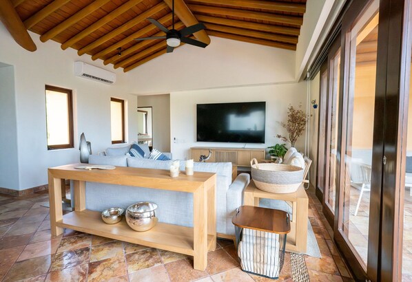 A modern living room with a wooden ceiling, a flat-screen TV, a sofa, a coffee table, and a large window. - Jaguar. Beach Facing House (10 steps) - Bucerías (Bucerías)