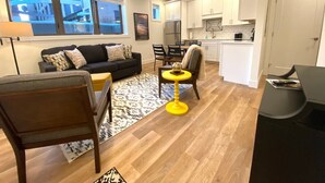 A modern living room with a black sofa, wooden chairs, a yellow table, and a kitchen area with white cabinets and a refrigerator.