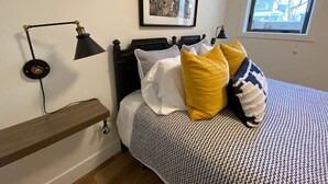 A bedroom with a bed, a wall-mounted lamp, a framed picture, and a window.
