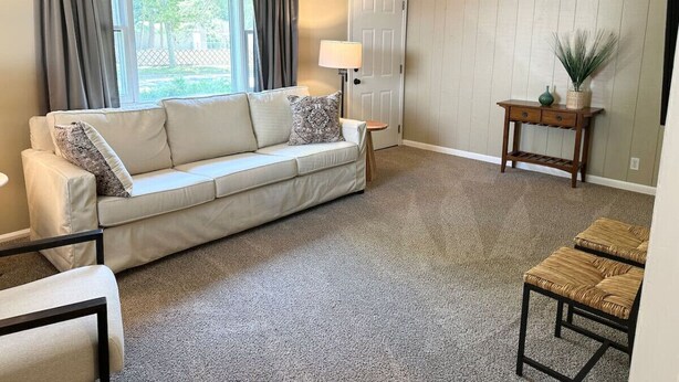 A living room with a beige sofa, a small table with a lamp, and a view of the outdoors through a window with curtains. - Cute and Cozy Near Parks (Midland)