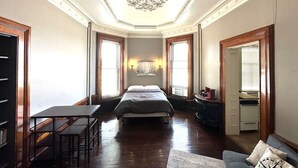 A spacious bedroom with a large bed, a desk, and a chair. There is a chandelier on the ceiling and a window with curtains. - Historical Studio Apt - Walk to All of Downtown (Bay City)