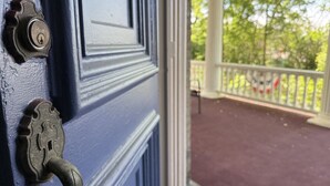 Una puerta azul con un ojo de cerradura y un pomo metálico, que da a un porche con barandilla y vistas a árboles.