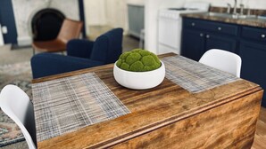 Una mesa de comedor con un centro de musgo verde, individuales a cuadros y sillas blancas en una cocina moderna con armarios oscuros y un horno empotrado.