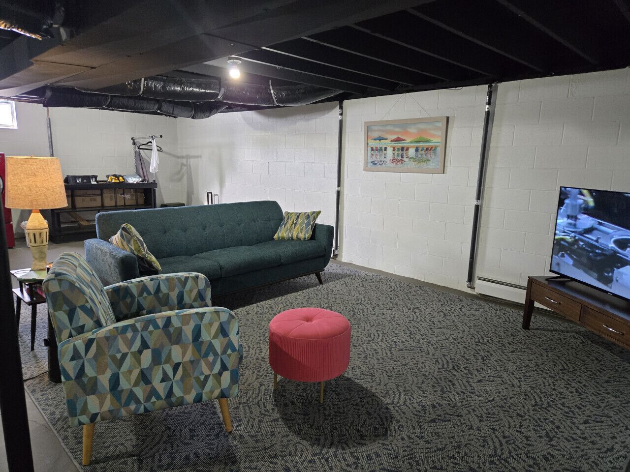 Un salon avec un canapé vert, un fauteuil à motifs et un pouf, une télévision et un tableau accroché au mur.