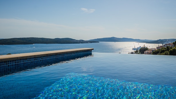 On aperçoit clairement une piscine, avec une terrasse en bois, et en arrière-plan, la mer et des collines.
