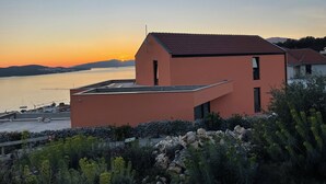 Un bâtiment moderne doté d’un toit plat, situé près d’une étendue d’eau, avec un coucher de soleil en arrière-plan.