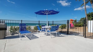An outdoor patio with blue chairs and a table, a blue umbrella, and a view of the beach and ocean.