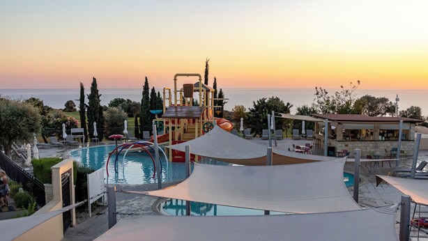 Un espace piscine doté d’un toboggan, d’une aire de jeux et offrant une vue sur la mer au coucher du soleil.