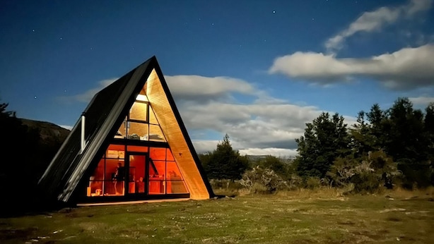 A modern, triangular-shaped building with a glass facade, situated in a grassy field with trees in the background. - Outscape Cerro Castillo (Río Ibáñez)