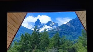 A view through a wooden-framed window of a mountainous landscape with snow-capped peaks and dense green forest. - Outscape Cerro Castillo (Río Ibáñez)