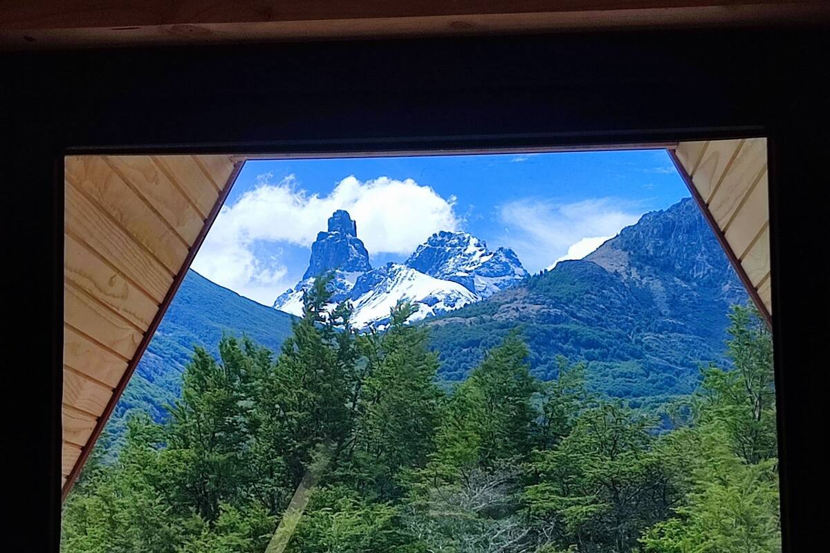 A view through a wooden-framed window of a mountainous landscape with snow-capped peaks and dense green forest.